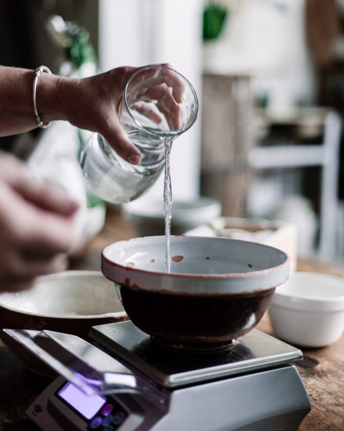 A bowl sits on top of a digital scale as a hand pours water from a jug into the bowl.