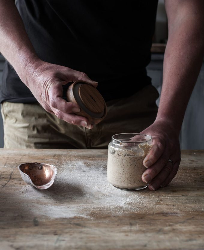 Bubbly sourdough starter in a glass jar on a wooden table. A mans hand is holding on to the lid of the jar.