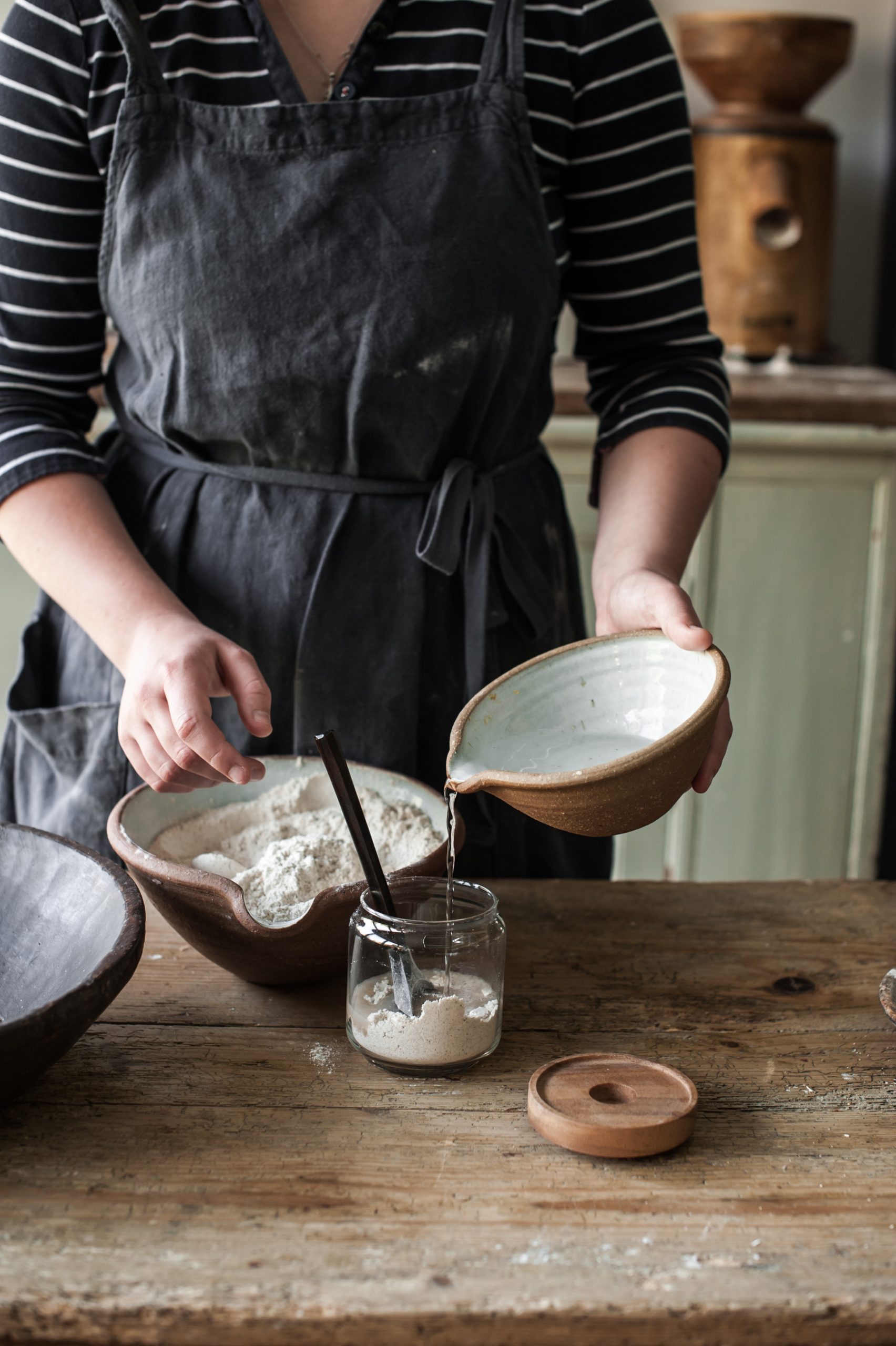 A woman with a bowl of flour and a smaller bowl with water, pouring this into a glass jar for a home for a starter.