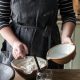 A woman with a bowl of flour and a smaller bowl with water, pouring this into a glass jar for a home for a starter.